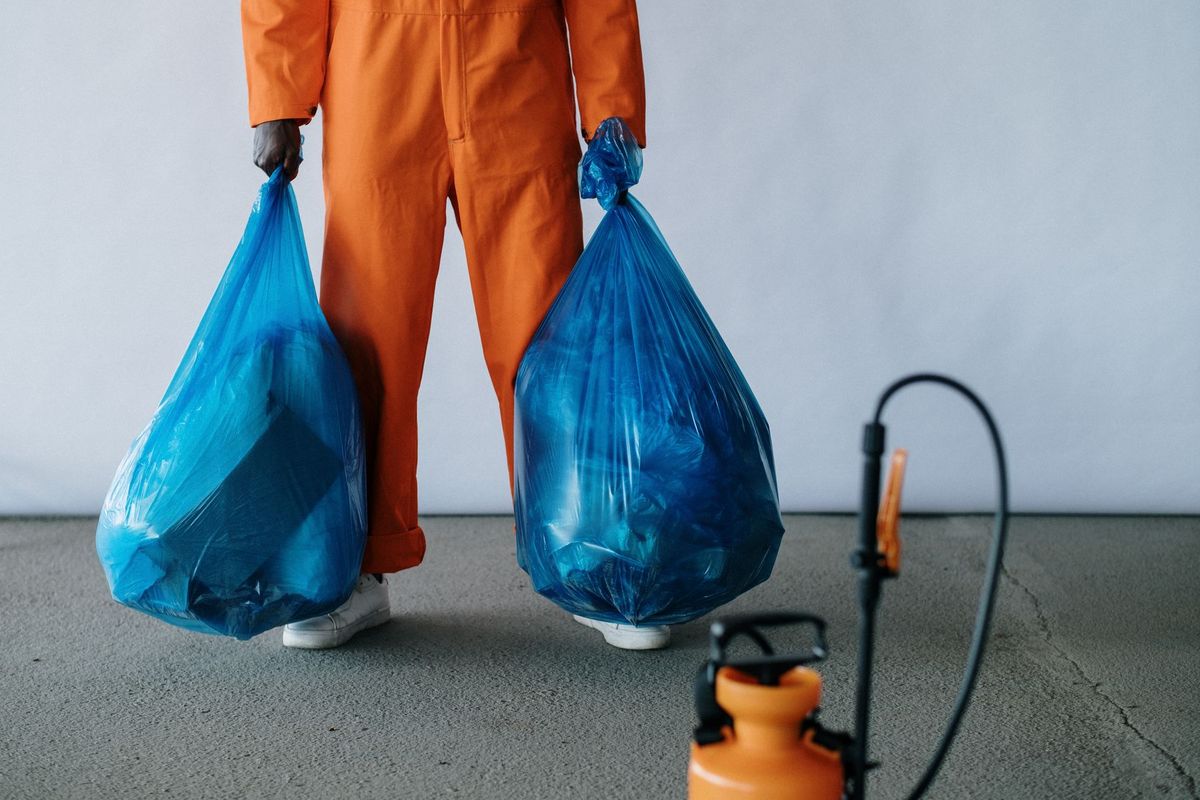Person in orange coveralls carrying two blue trash bags next to a pump sprayer