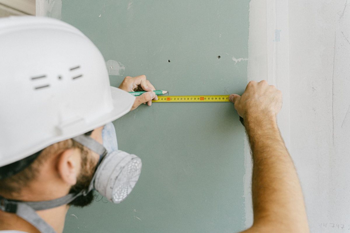Worker in a hard hat and respirator measuring along a wall with a tape measure