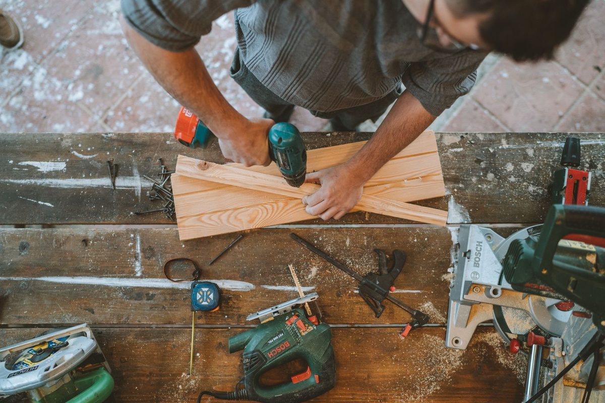 A person cutting a wooden board on a cluttered workbench with power tools