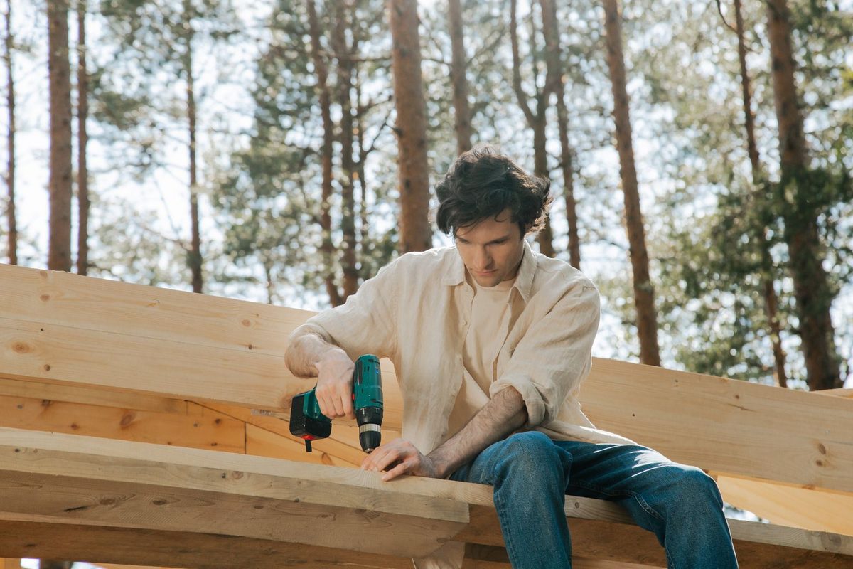 A man sitting on stacked lumber outdoors, driving a screw into a board with a cordless drill