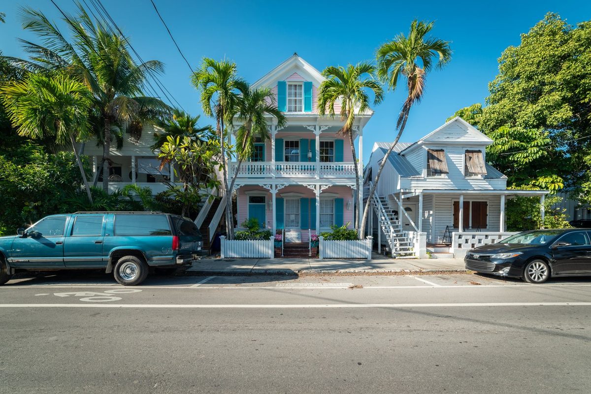 A row of older two-story wood-frame houses with porches and shutters behind palm trees