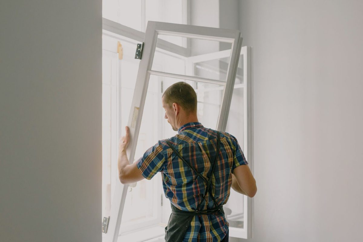 A worker in a plaid shirt lifting a new white window frame into an opening
