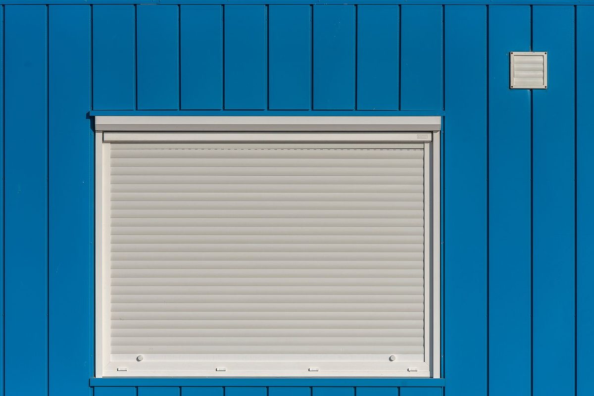 White roll-down storm shutter closed over a window on a blue exterior wall
